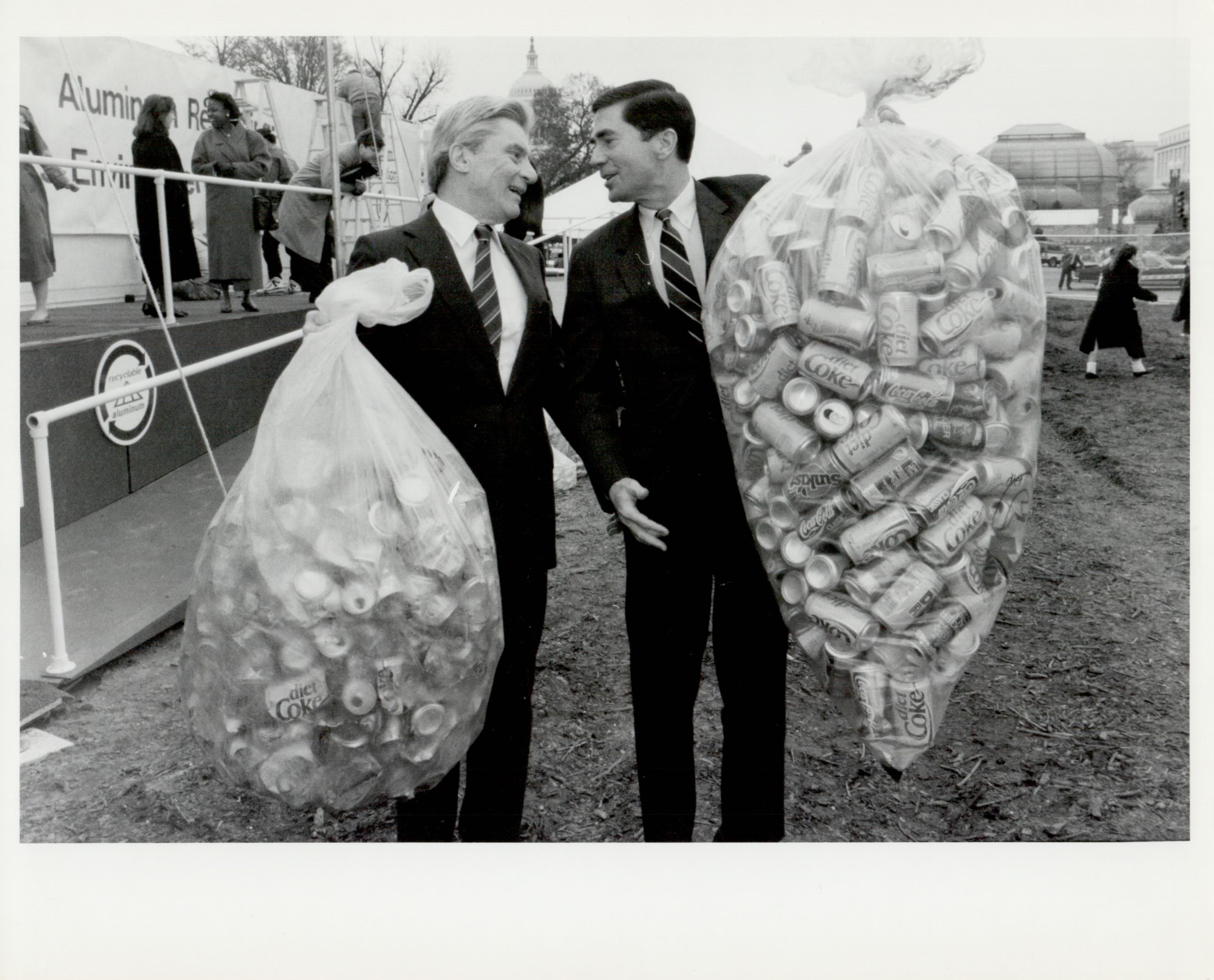 Photo of two men in suits carrying clear plastic bags full of aluminum cans to recycle